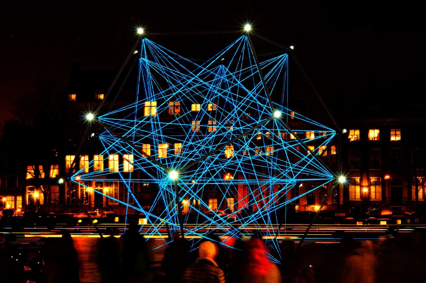 A huge star-shaped sculpture of glowing blue cords in front of canal houses with warm lit windows at night, with boat light trails on the water and blurred visitors in the foreground