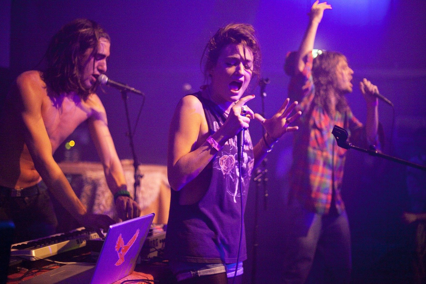 A band performing in violet light at the Westergasfabriek, the singer belting into the microphone with her eyes closed, a shirtless keyboard player at a synth and laptop, another singer with an arm raised