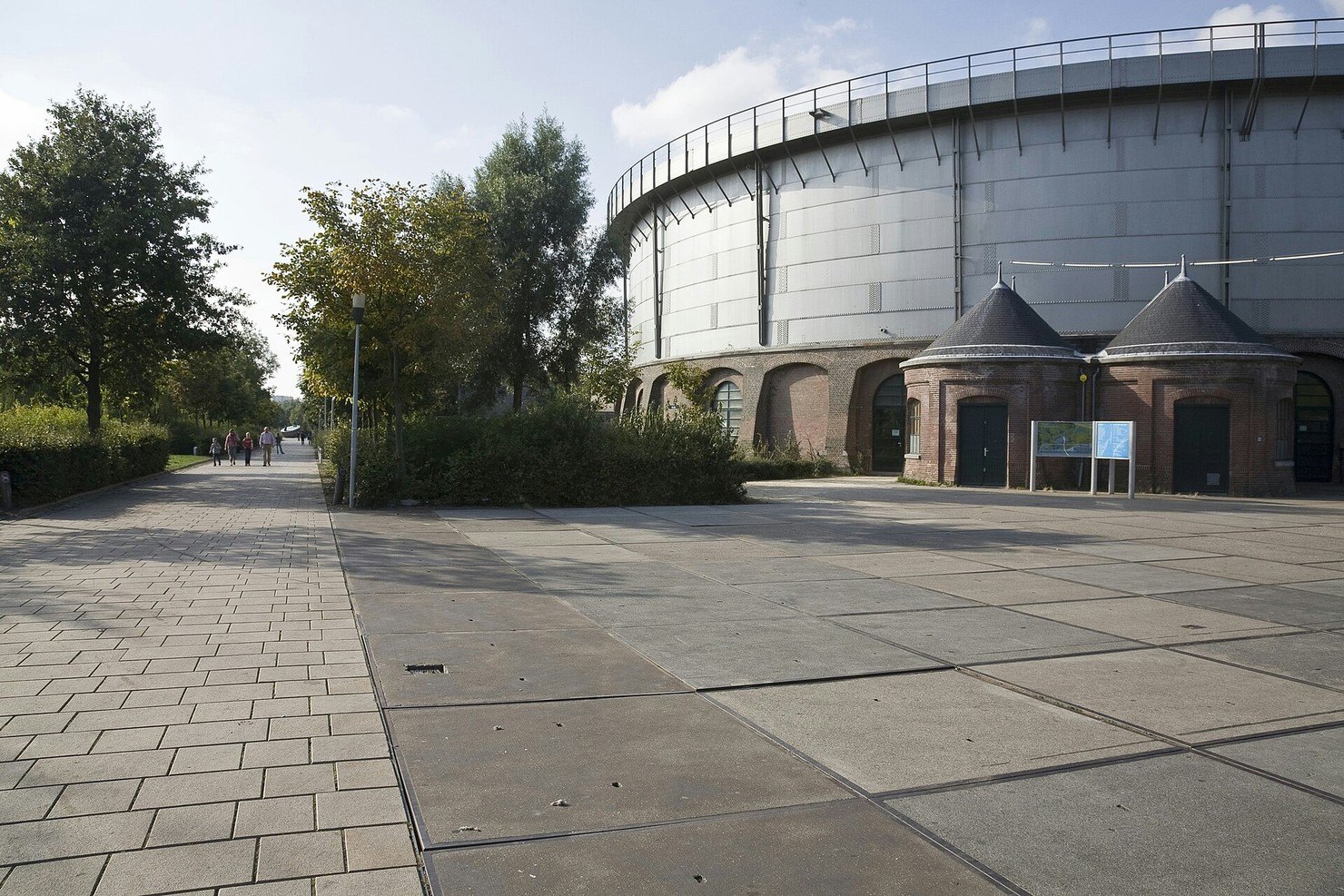 The round iron gasholder of the Gashouder at Westergas in Amsterdam, with two small brick towers at its base, seen across a paved square on a sunny day