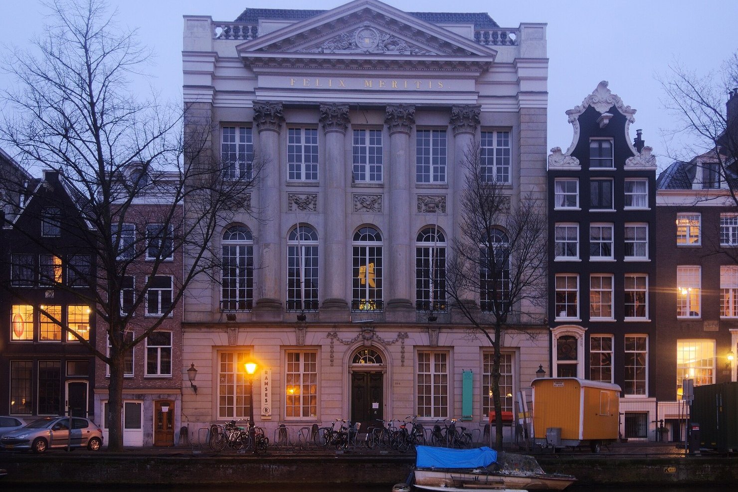 The neoclassical facade of Felix Meritis on the Keizersgracht in Amsterdam at dusk, with FELIX MERITIS lettered under the pediment, bicycles along the railings and the canal in front