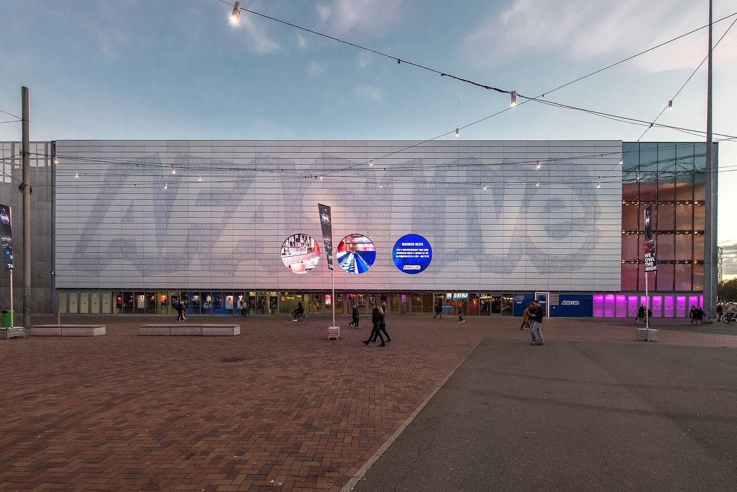 The pale grey facade of AFAS Live in Amsterdam at dusk, with the venue name printed large across it, festoon lights strung over a brick square and a few people walking past