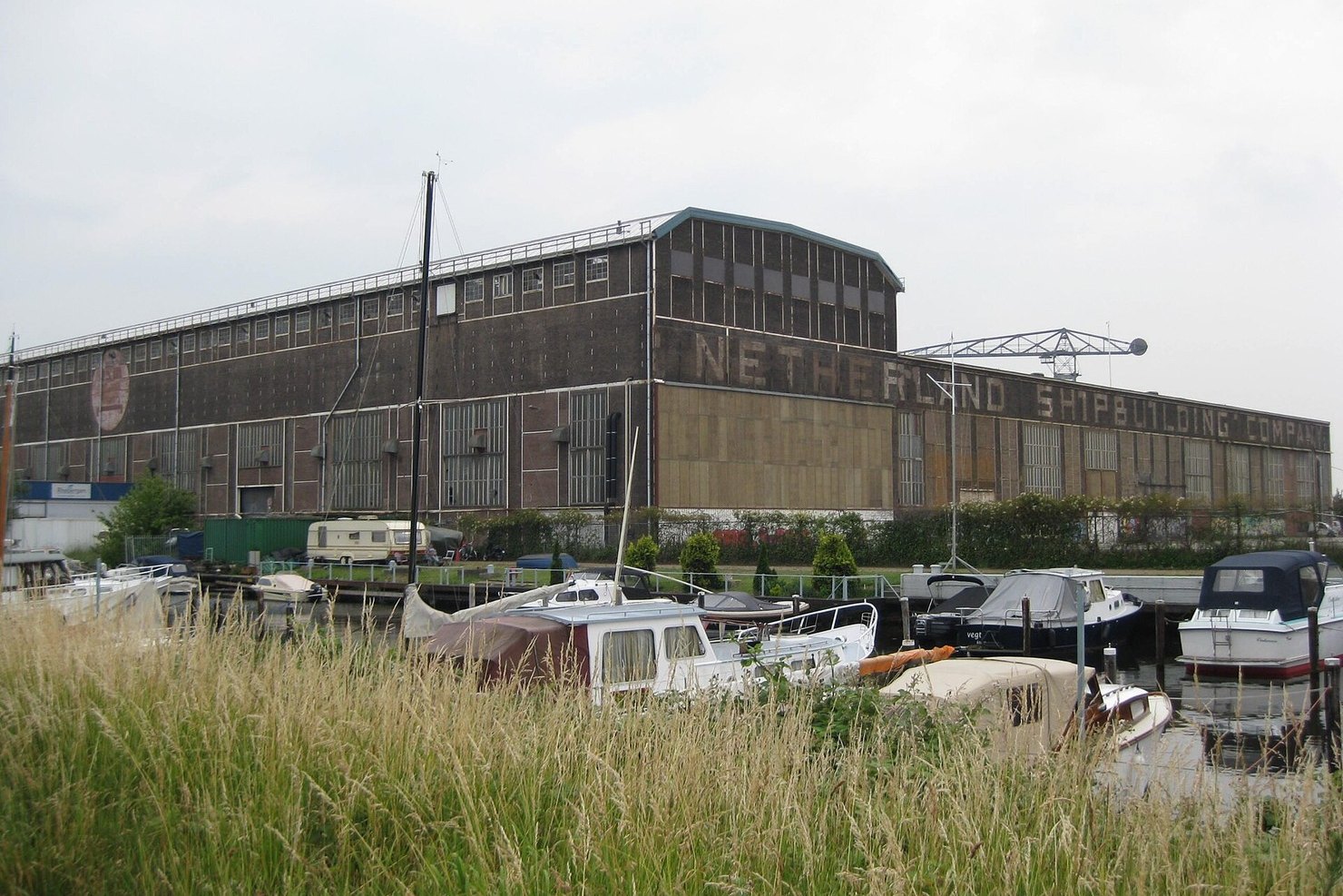 The long dark shipbuilding hall at the NDSM wharf in Amsterdam-Noord, with Netherland Shipbuilding Company lettered along its side and small boats moored in front