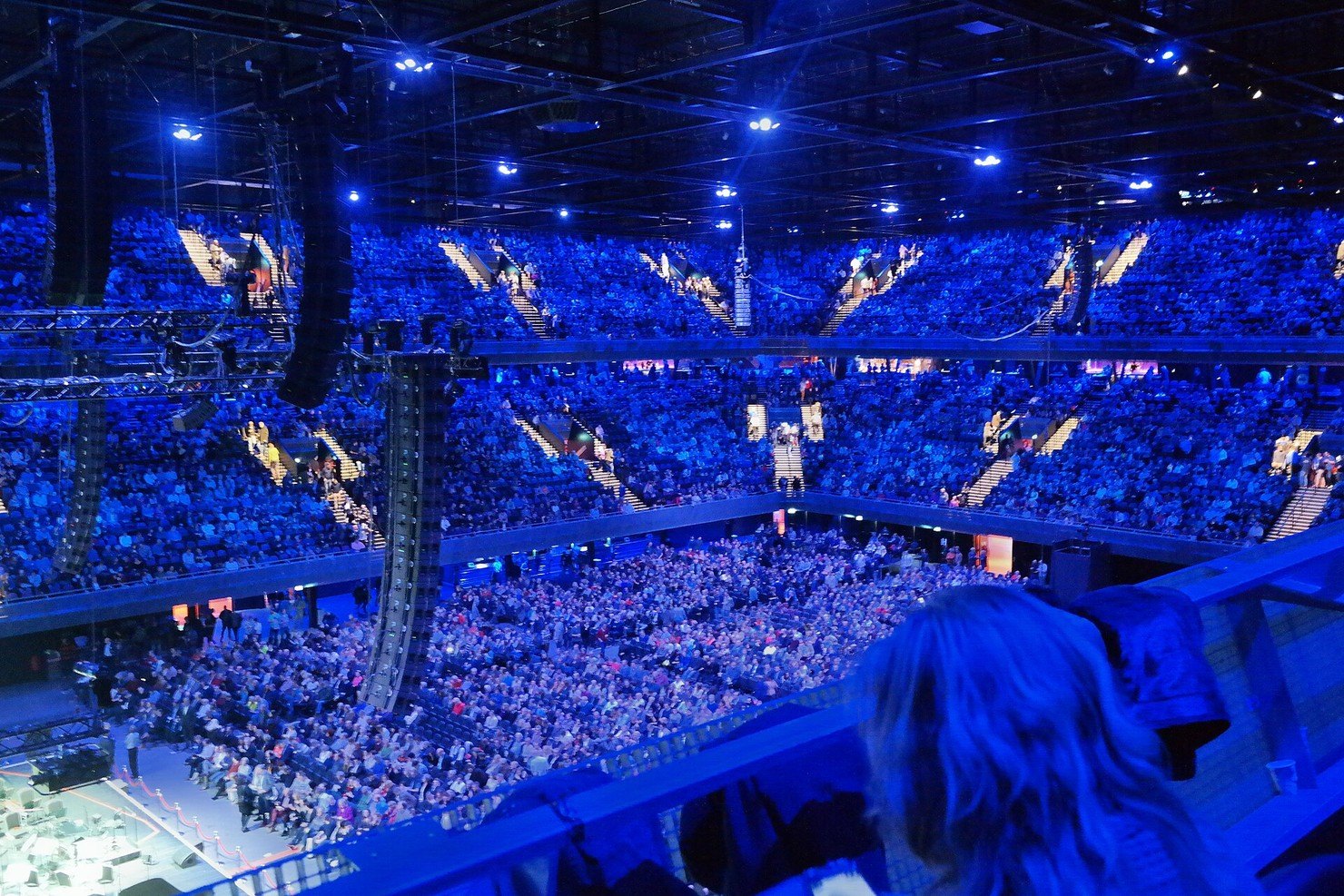 Inside the Ziggo Dome in Amsterdam Zuidoost, seen from the top tier: the floor and two rings of stands full under blue light, with speaker stacks hanging from the roof