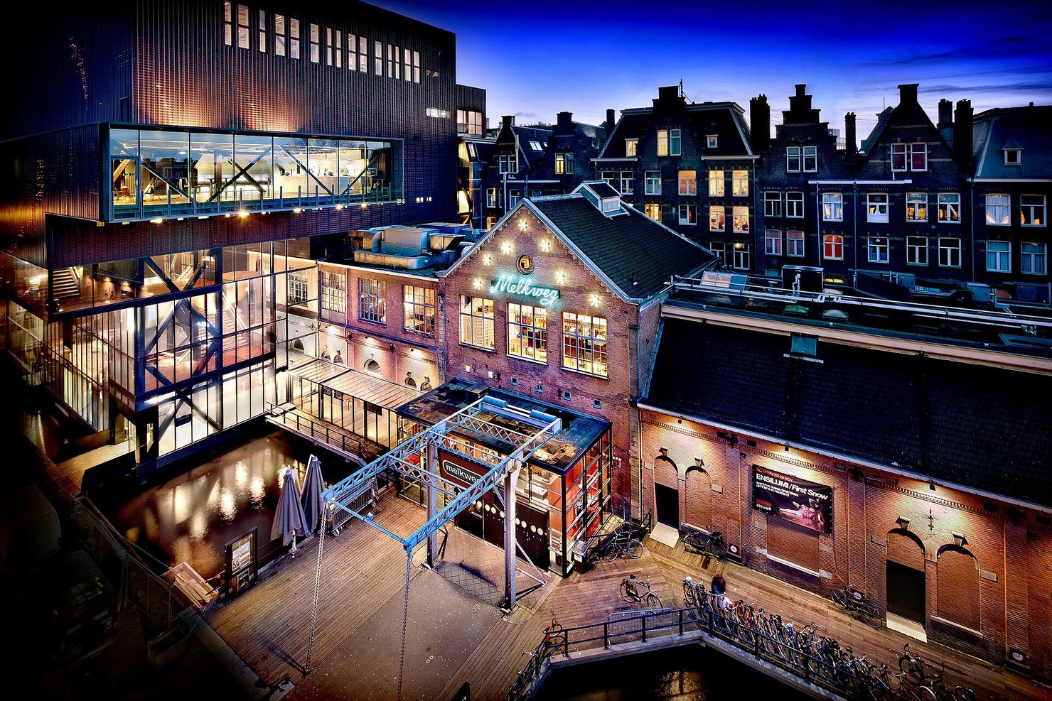 Melkweg in Amsterdam at dusk, seen from above: the neon Melkweg sign on the old brick building, the glass and steel extension beside it and bicycles parked on the canal-side deck