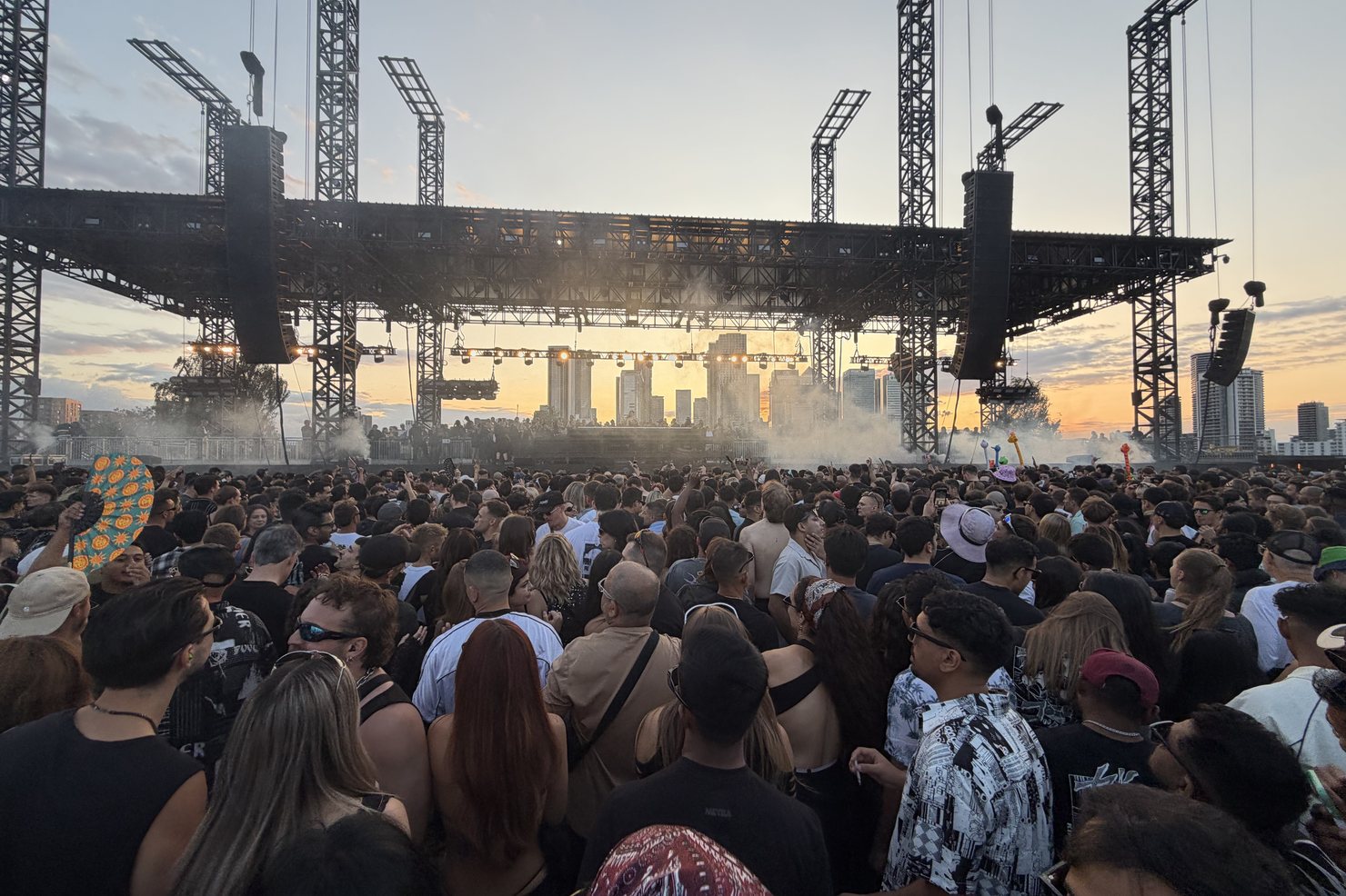 Golden hour at Magazine Open-Air: the open-air stage rig with hanging line arrays, the Canary Wharf skyline behind the decks and thousands of people on the field, Greenwich Peninsula, 21 August 2026.