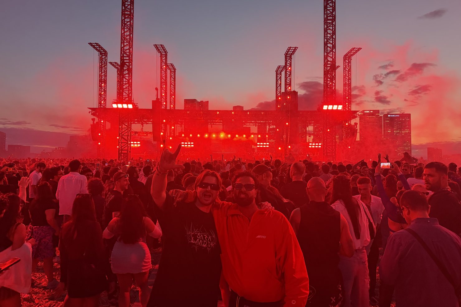 The stage and rig towers washed in red light at dusk during the hard techno night at Magazine Open-Air, smoke drifting over the crowd, the Canary Wharf towers visible at the right.