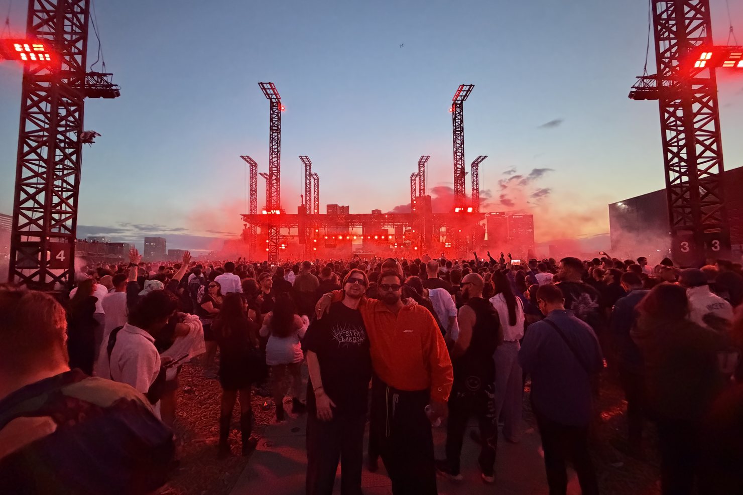 The Magazine Open-Air field at dusk between numbered rig towers, red light over the distant stage and wood chips covering the ground in the foreground.
