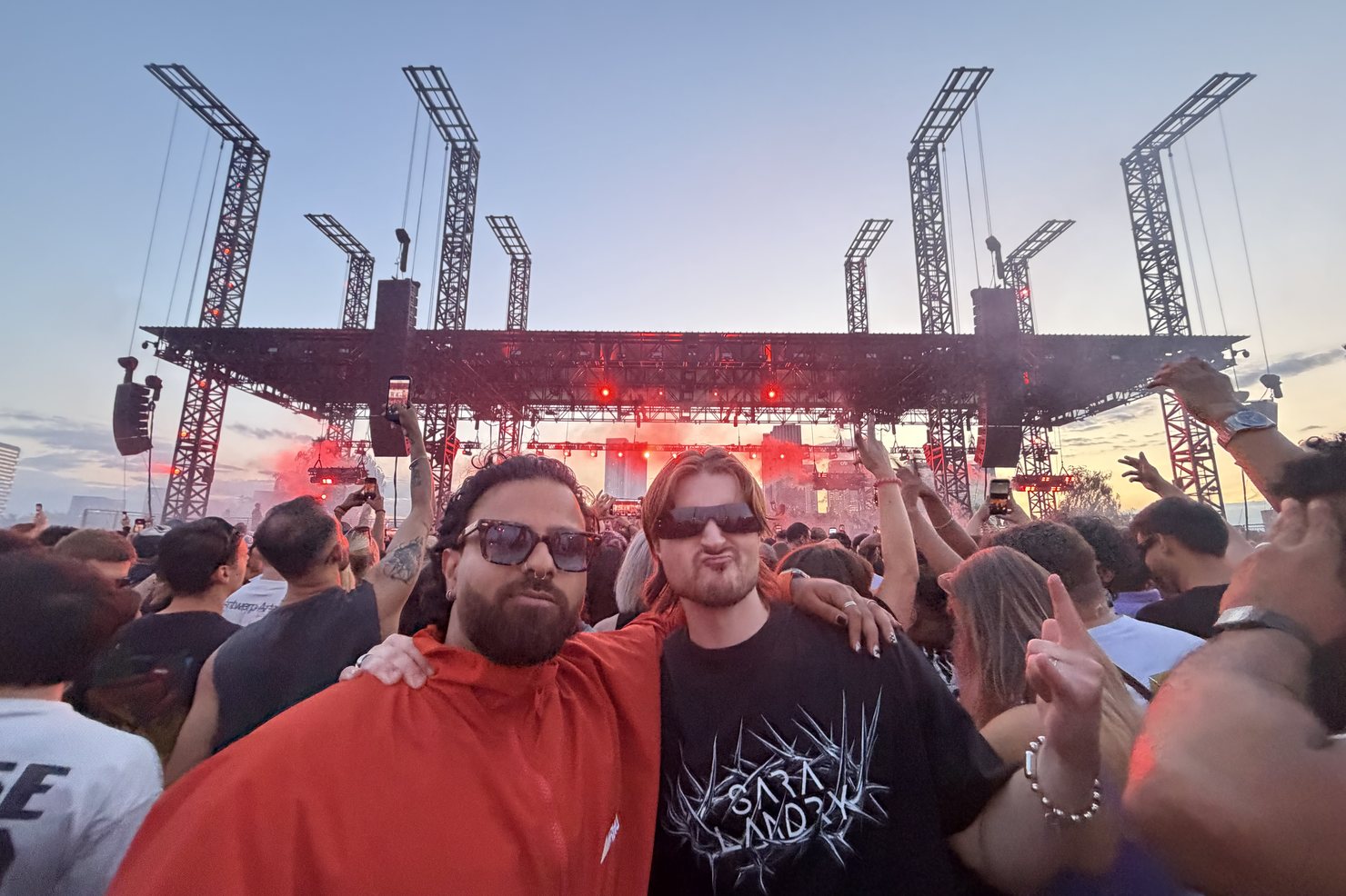 Ravi Sharma, founder of Neon Black, in a red jacket with CABBA LAGA in a spiked Sara Landry T-shirt in front of the Magazine Open-Air stage at sunset, phones raised across the crowd behind them.