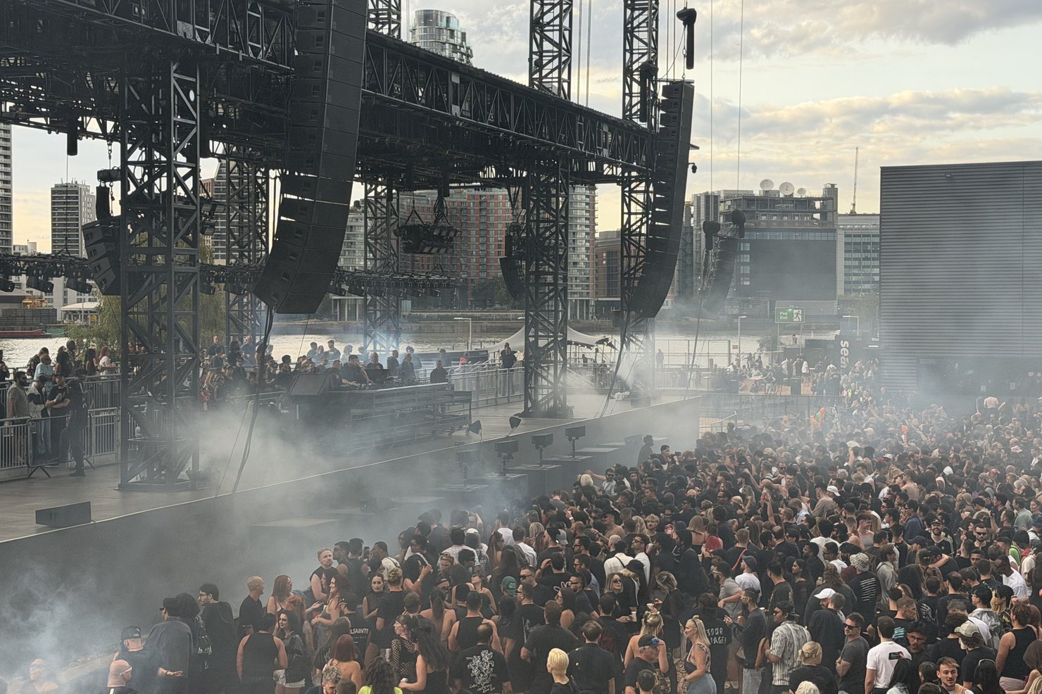 The stage seen side-on from the elevated viewpoint at Magazine Open-Air, line arrays and smoke over the deck, the crowd below and the Thames with riverside towers behind.