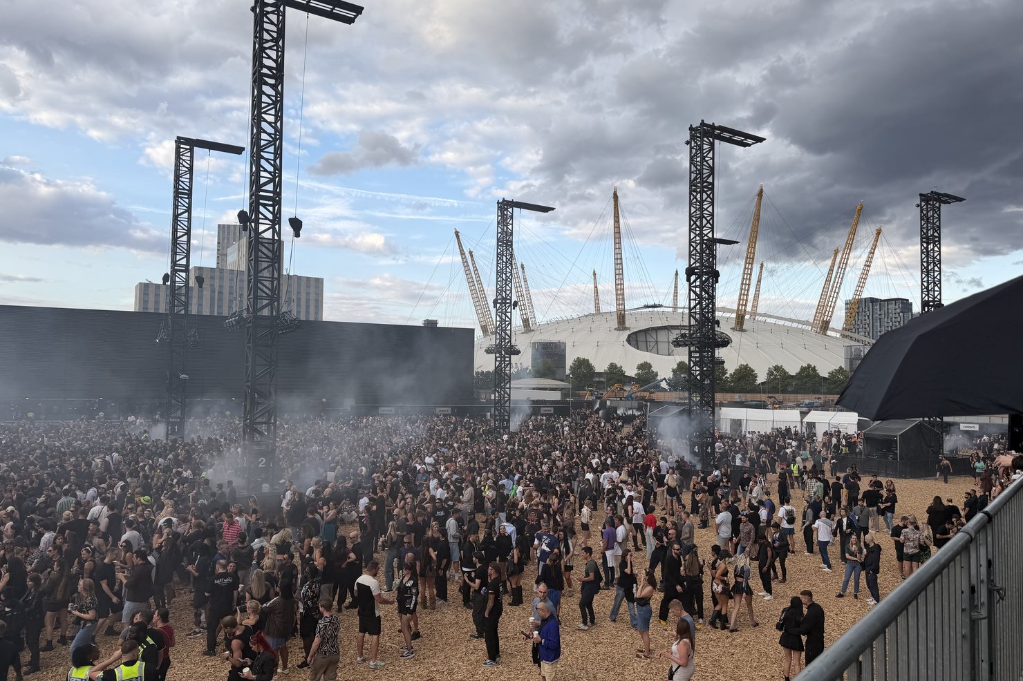 The Magazine Open-Air field from above with wood chip covering the ground, numbered rig towers, drinks stalls and the yellow masts of The O2 dome directly behind the site under heavy clouds.