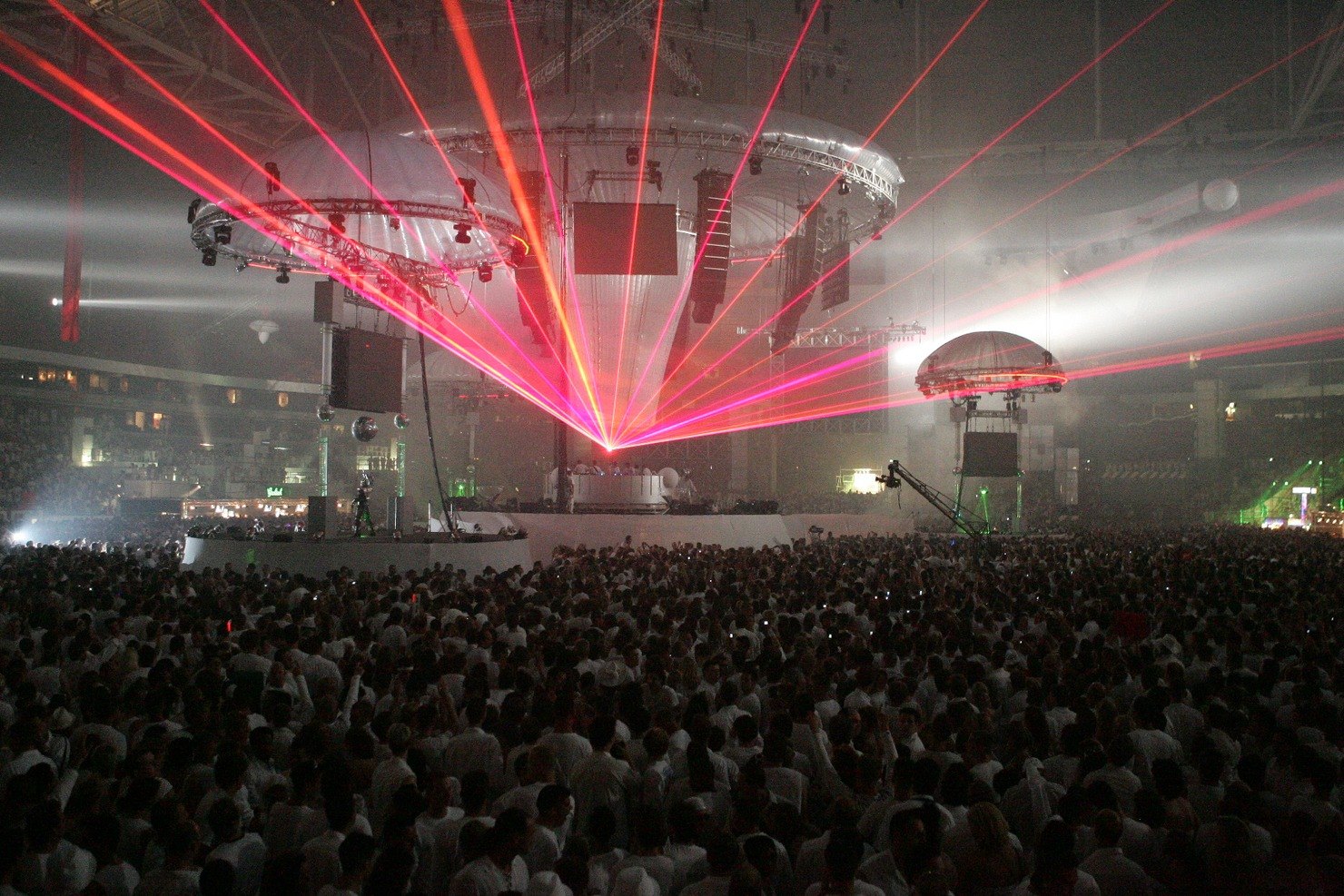 Red and pink laser beams fan out from the DJ booth beneath domed stage rigs over a packed crowd dressed in white in the Amsterdam ArenA