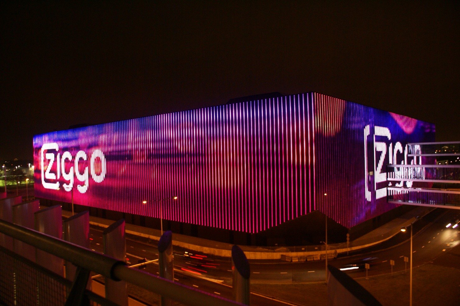 The Ziggo Dome's LED facade glows pink and purple at night above a road streaked with car light trails, seen from a car park deck in Amsterdam Zuidoost