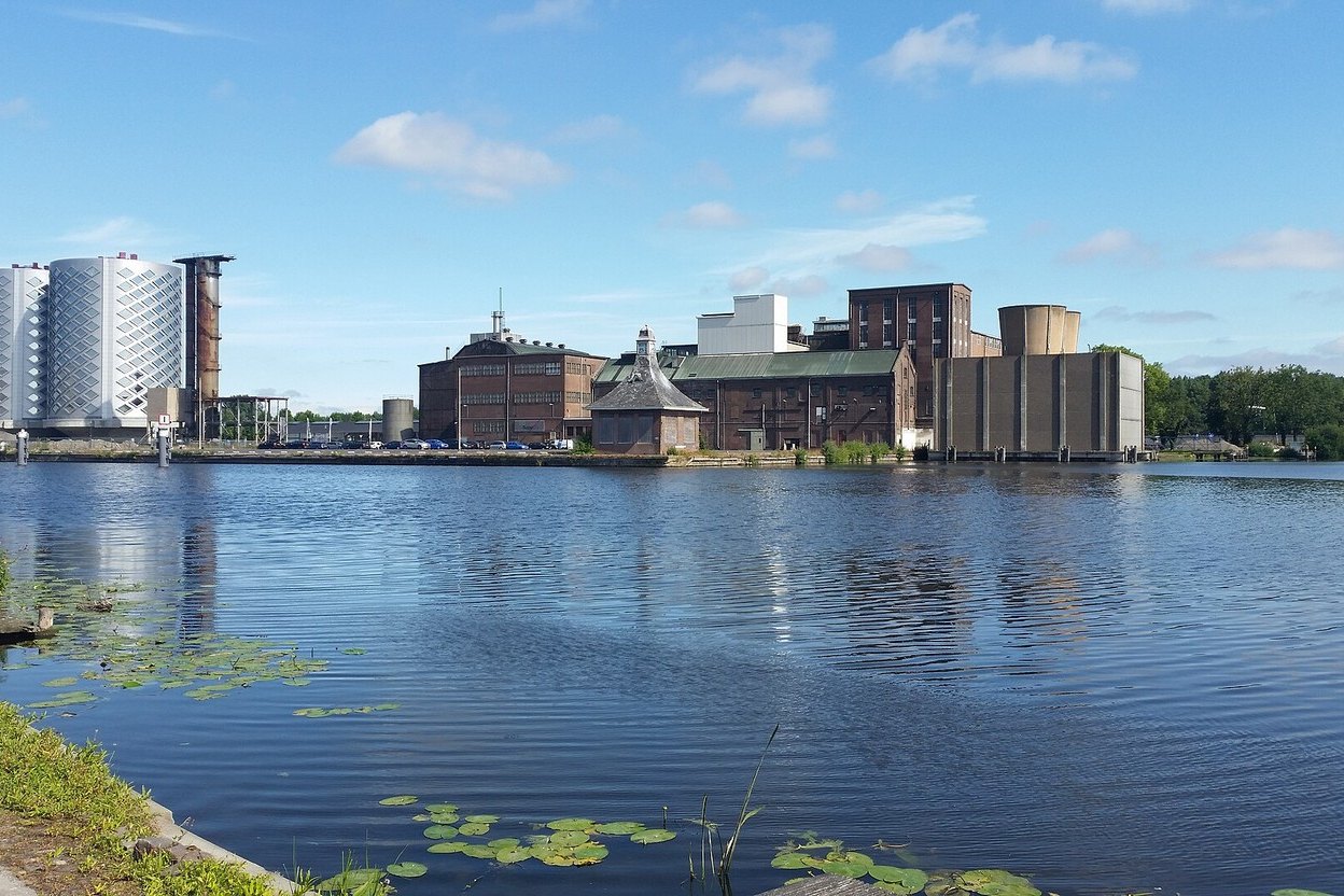 The former sugar factory complex at SugarCity in Halfweg, brick factory halls and two tall lattice-clad silos, seen across calm water with lily pads under a blue sky