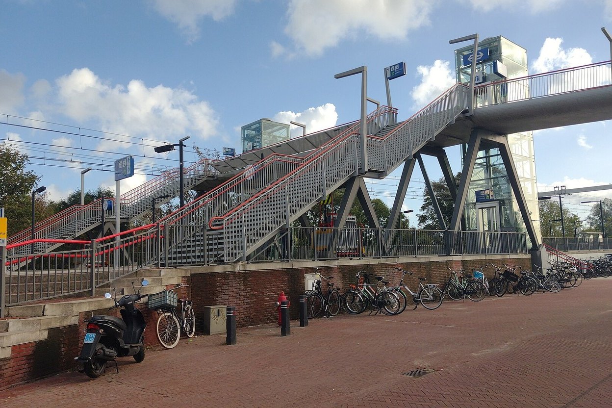 The footbridge, stairs and glass lift towers of Halfweg-Zwanenburg station, with bikes parked along a brick wall below, on a sunny day