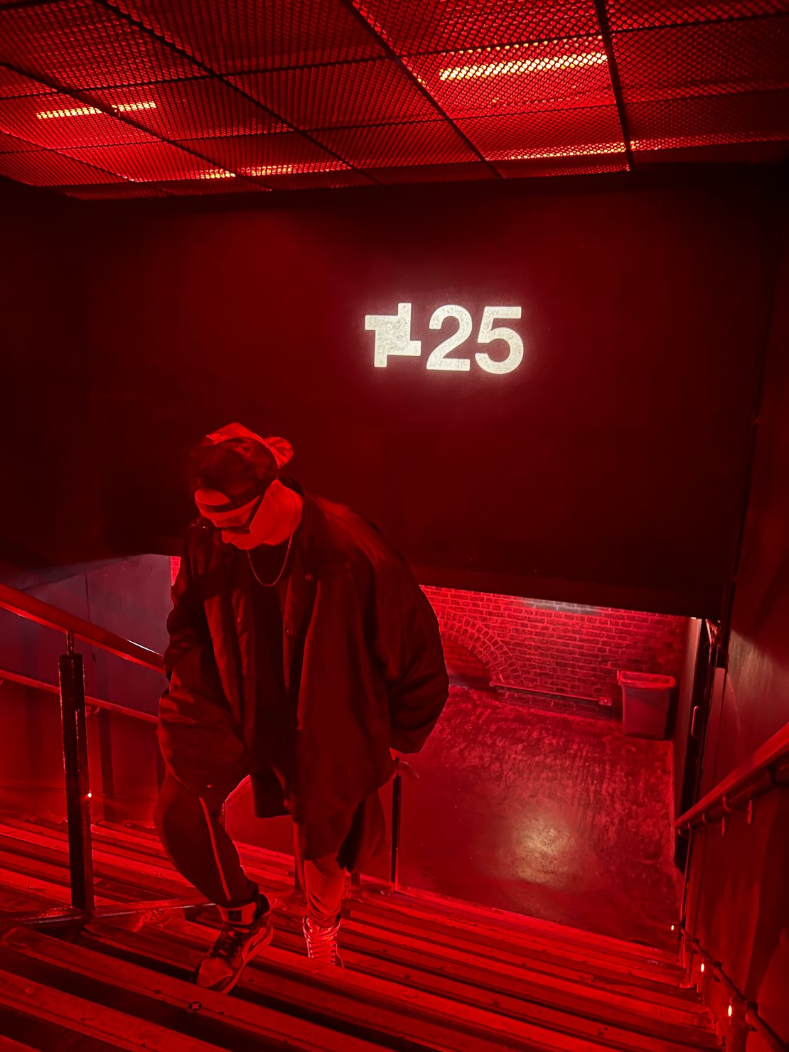 MNEEMO (Yaroslav Gorovoy) in a jacket descends red-lit stairs with "25" sign above. Industrial setting with brick walls and intense lighting. 25 year anniversary of fabric London. 
