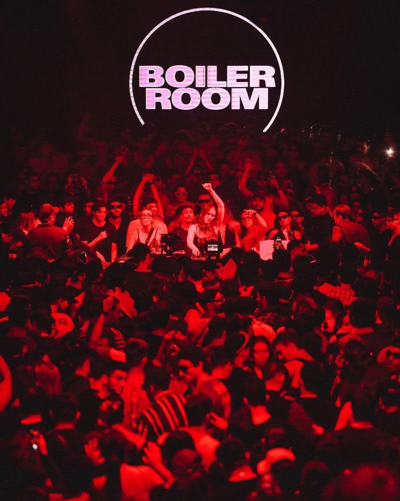 Boiler Room event photo with a packed red-lit crowd surrounding the DJ booth, with the Boiler Room logo projected above the dancefloor.