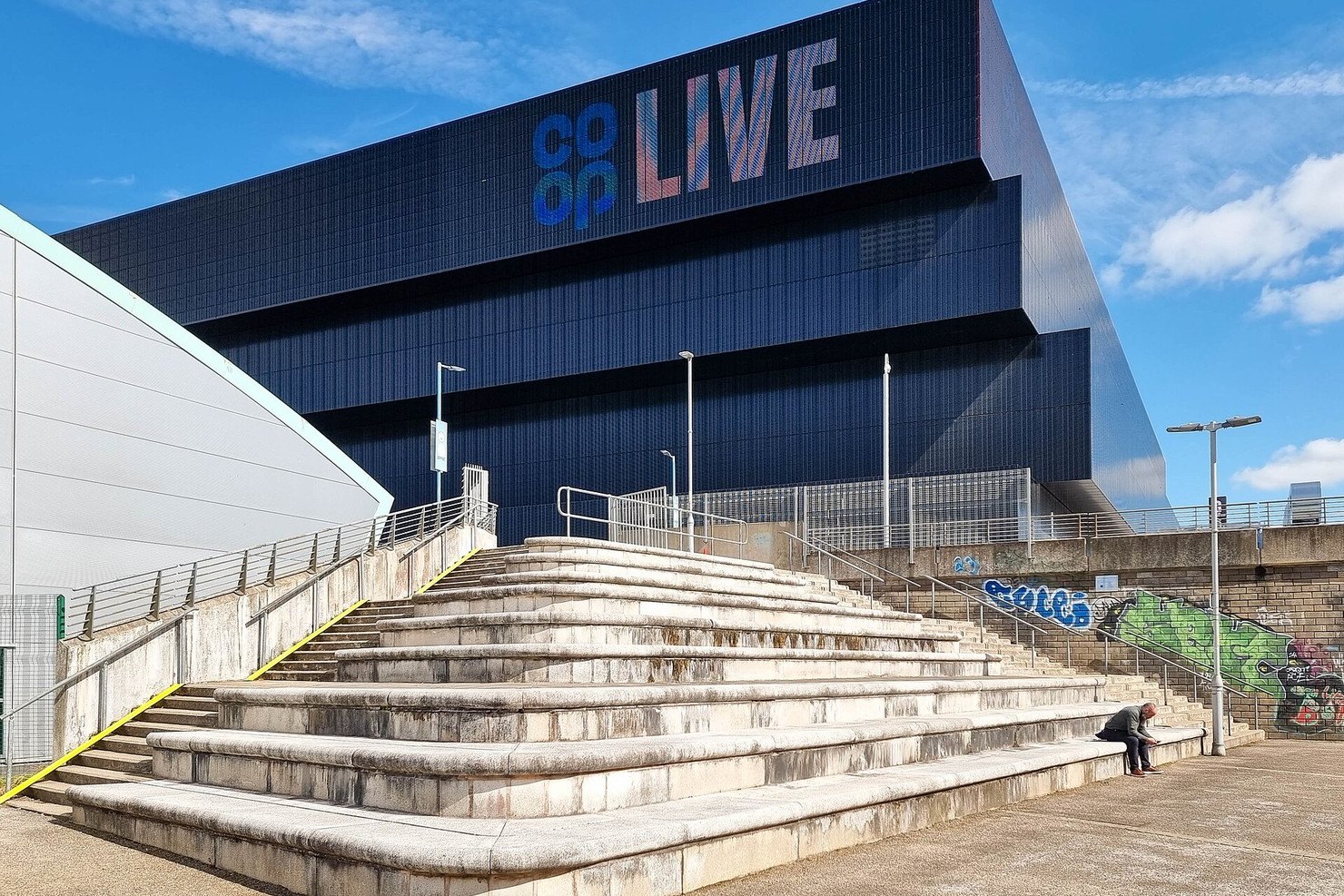 Co-op Live arena in Manchester in daylight, a dark tiled block with the Co-op Live sign lit in colour above wide stone steps