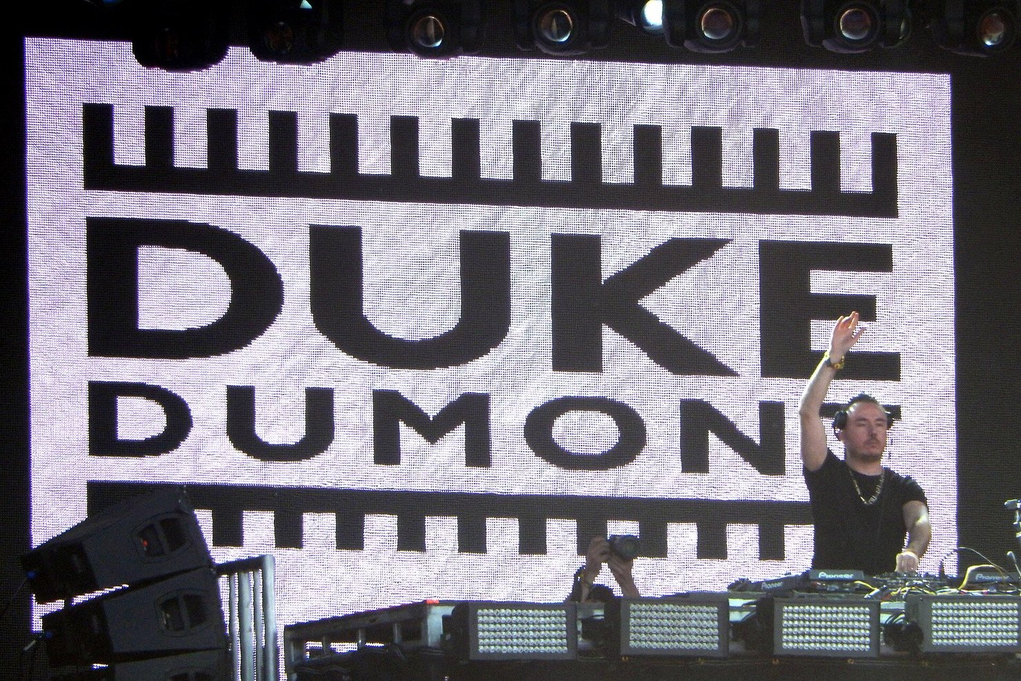 Duke Dumont DJing with one arm raised in front of a giant Duke Dumont logo screen at Lollapalooza Chicago, 2014