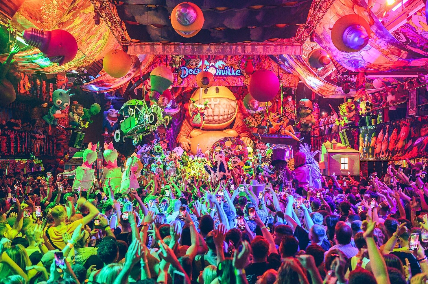 An elrow party in full flow: a crowd with hands and phones raised under inflatables, a giant smiling character and a Delusionville sign
