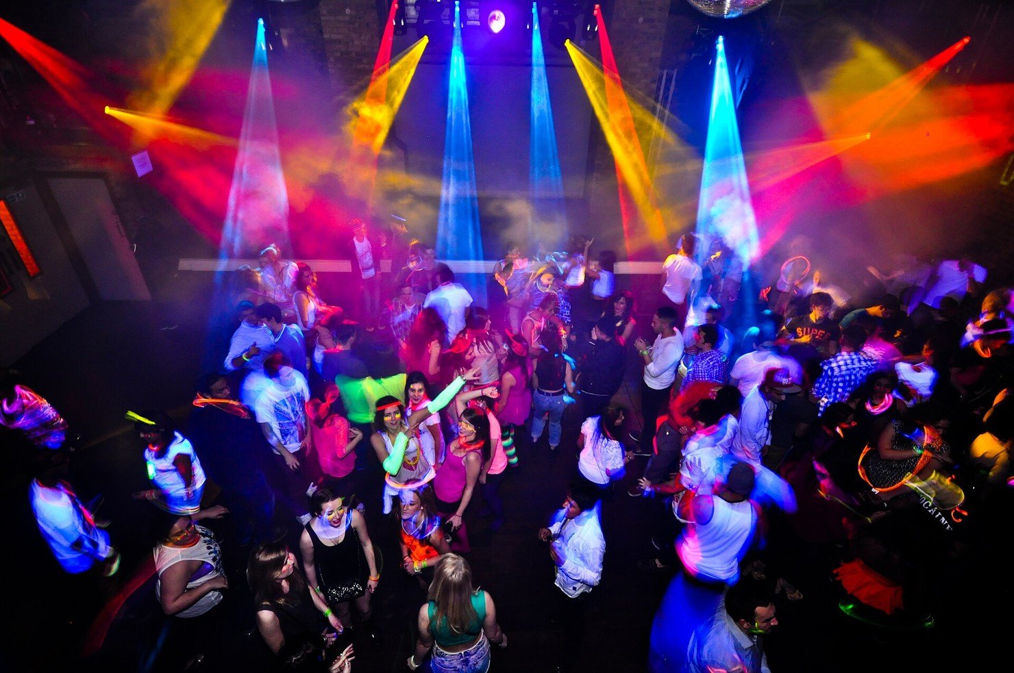 A packed fabric dancefloor seen from above, dancers under coloured moving-head lights and haze