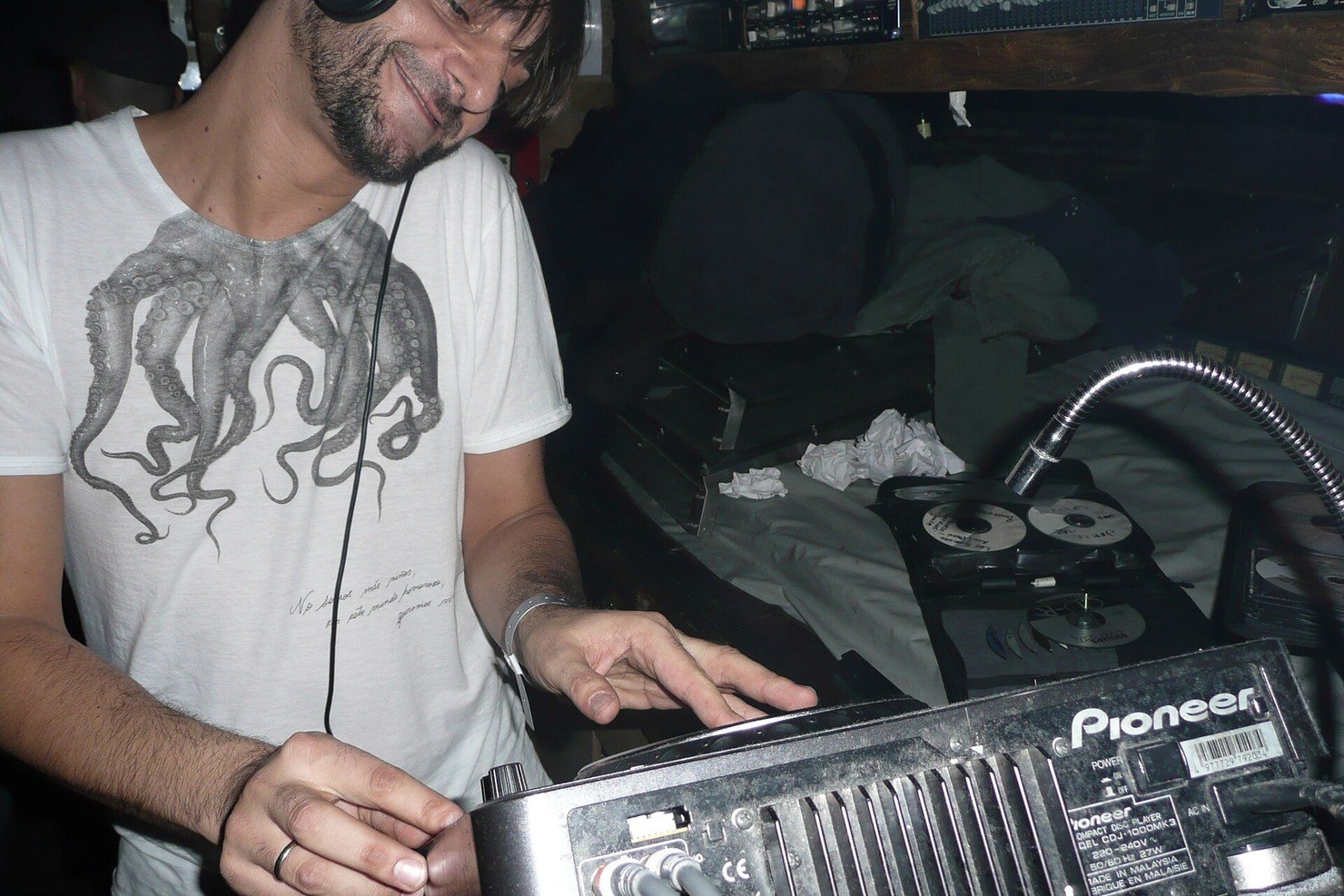 Ricardo Villalobos smiling in the fabric DJ booth as he adjusts a Pioneer CDJ, headphones round his neck