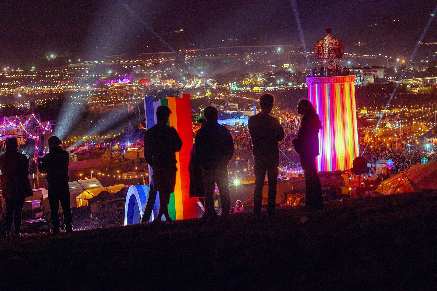 Glastonbury Festival at night from the hill, silhouettes looking down over the lit-up site and the Ribbon Tower