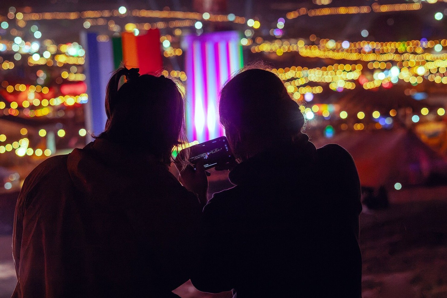 Two people silhouetted against the lights of Glastonbury Festival at night, looking down over the site from the hill