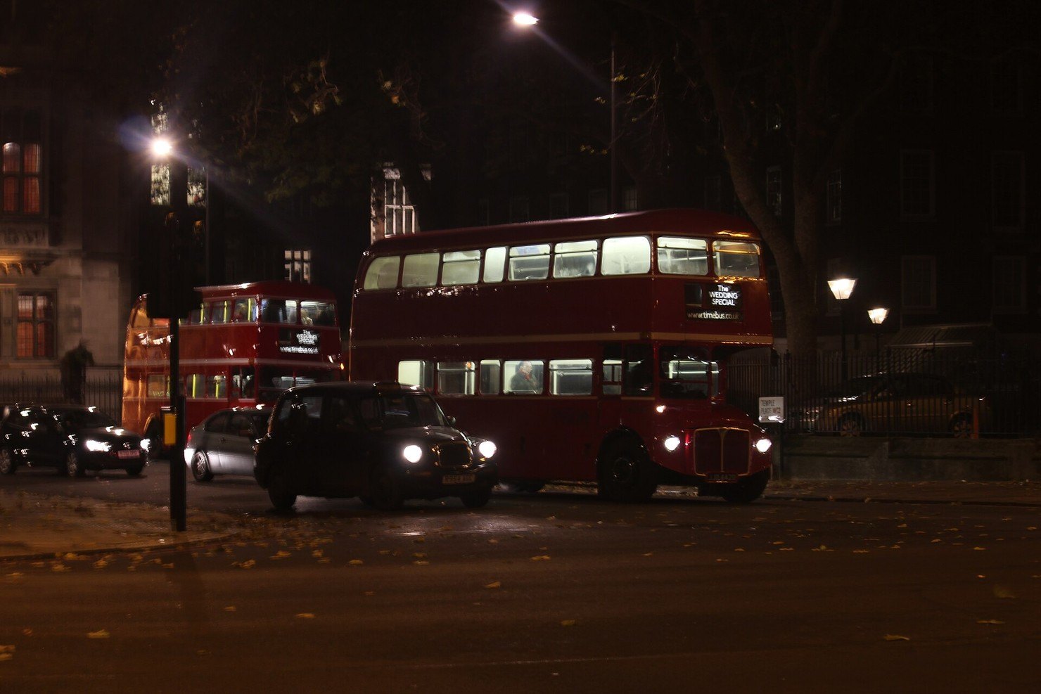 A black London taxi with its headlights on passing two red double-decker buses on a dark street lit by lamps