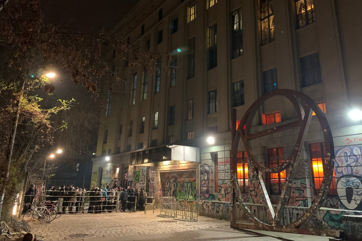The queue outside Berghain in Berlin at night, people waiting behind barriers beneath the club's concrete facade