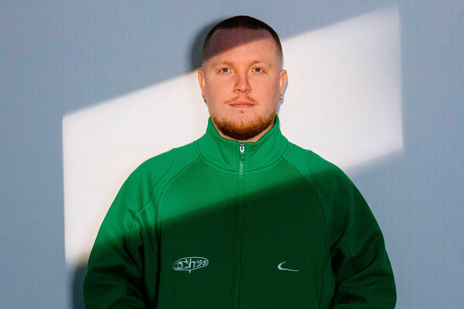 Chris Stussy in a green hoodie with logos, standing against a wall with a rectangular patch of sunlight. Calm expression.