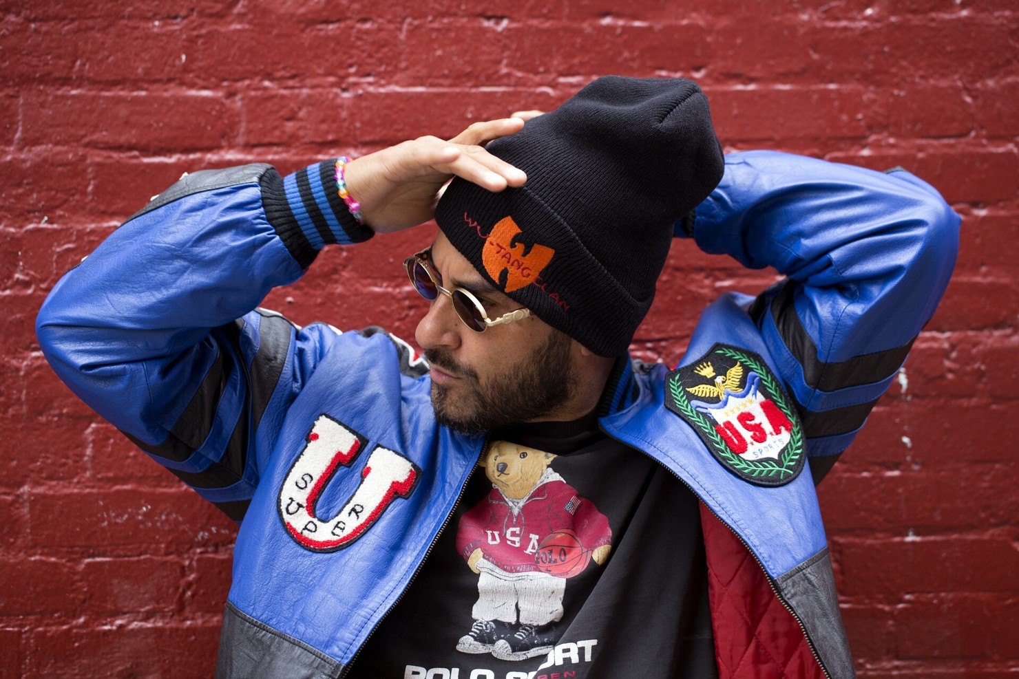 Armand Van Helden in round sunglasses, a black Wu-Tang Clan beanie and a blue leather varsity jacket, one hand on his head, against a red brick wall