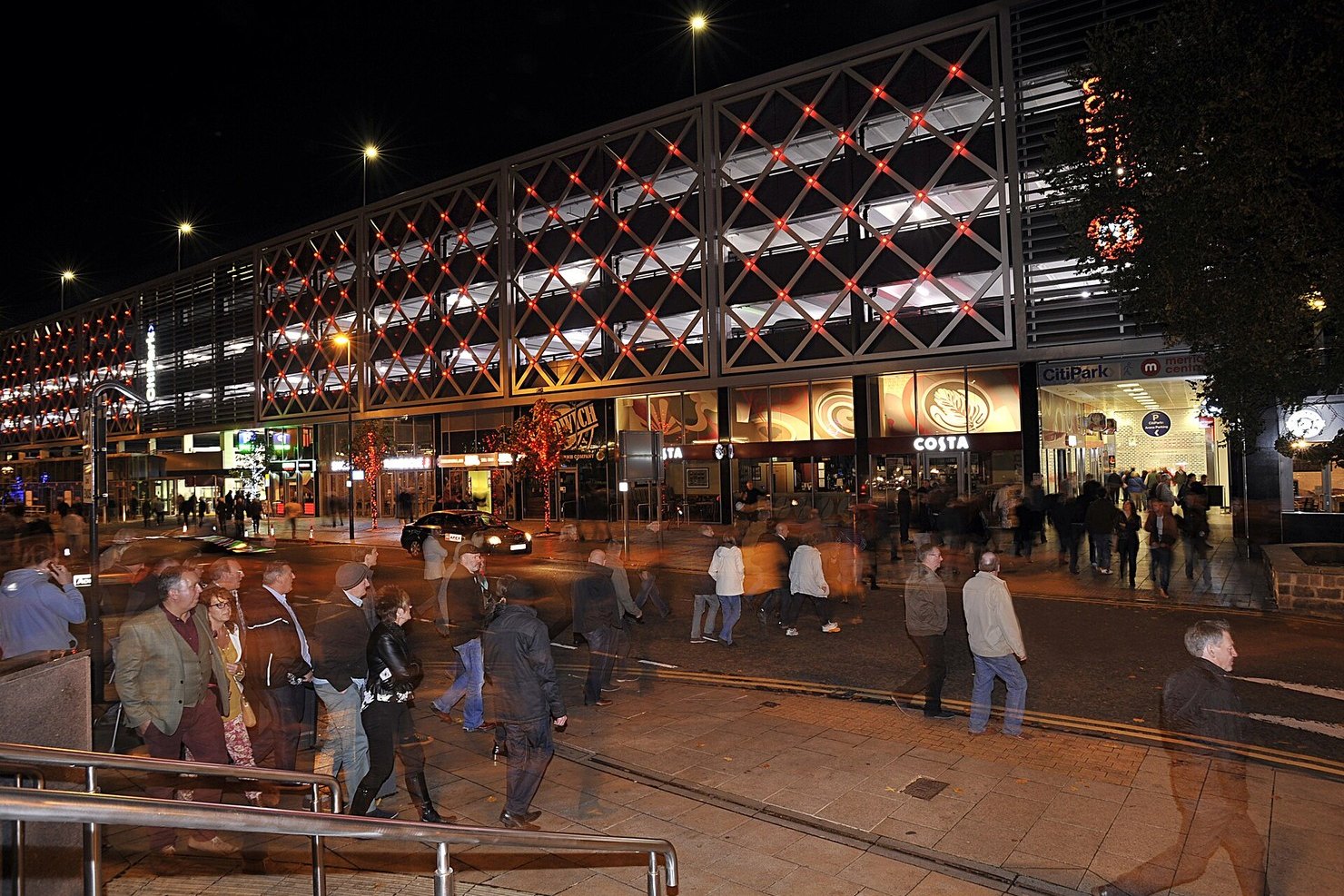 People walking past the lit front of the Merrion Centre on Merrion Way in Leeds at night, its lattice facade glowing red and white above the shopfronts