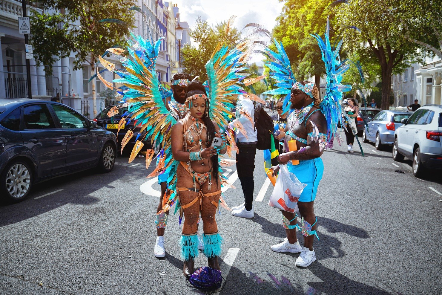 Three masquerade performers in feathered carnival costumes on a west London street before the Notting Hill Carnival parade