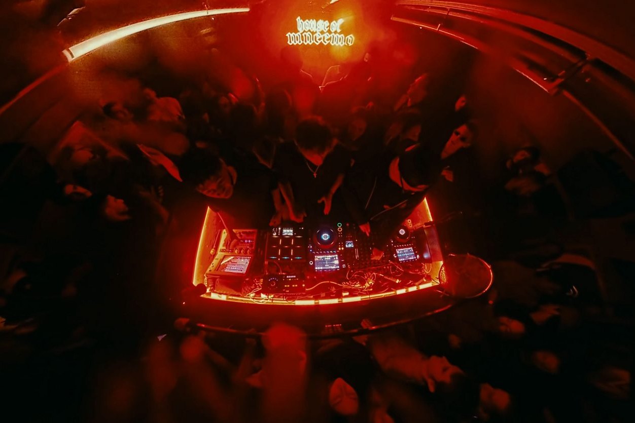 Overhead view of a DJ booth ringed in red light at a HOUSE OF MNEEMO night, the crowd pressed in around the decks