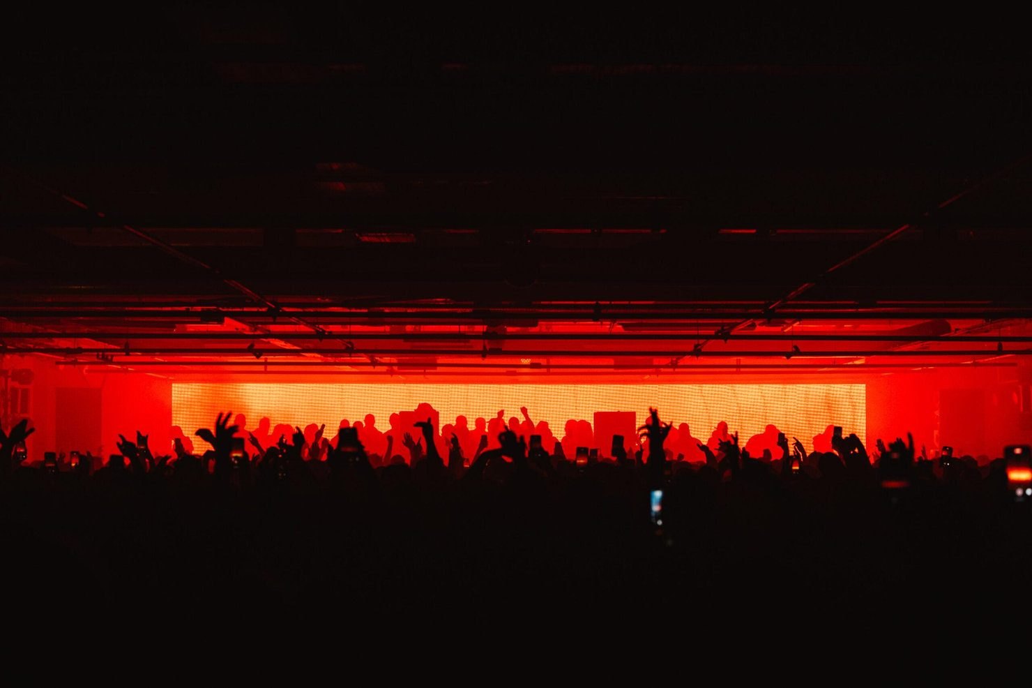 A crowd in silhouette against a wall of red light at Drumsheds in Tottenham, phones raised towards the stage