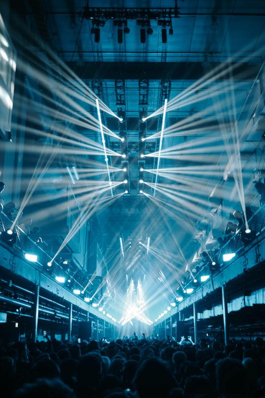 Printworks 2026 Concert scene with blue lights beaming across a crowded venue. People stand below the stage, beams creating an energetic atmosphere.