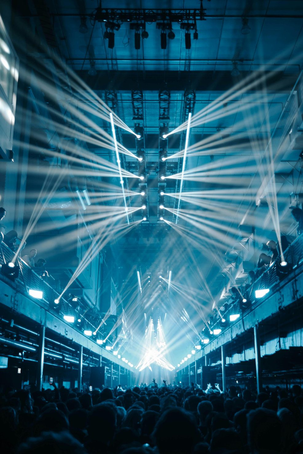 Printworks 2026 Concert scene with blue lights beaming across a crowded venue. People stand below the stage, beams creating an energetic atmosphere.