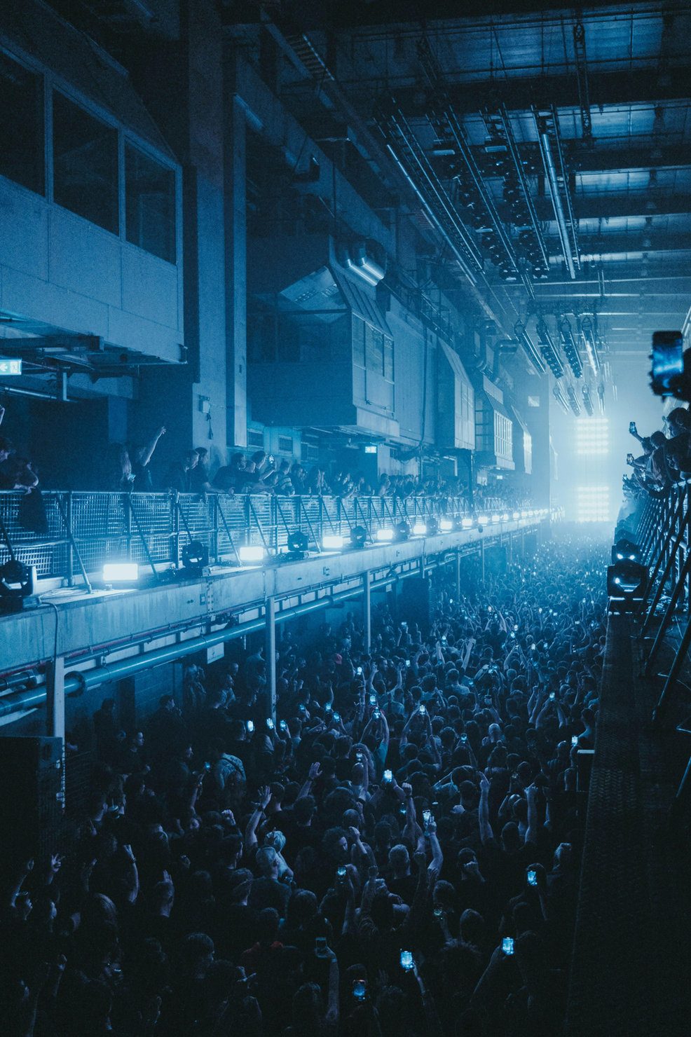 Printworks 2026 Concert crowd in a large, industrial-style venue with blue lighting. People raise phones, capturing the moment. Energetic atmosphere.
