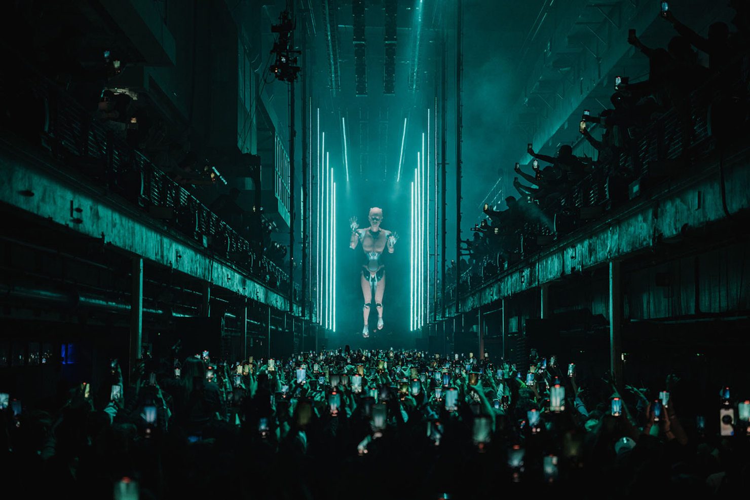 Printworks 2026 A futuristic concert scene with a large holographic figure on stage, surrounded by a crowd holding phones. Neon blue lights create a vibrant atmosphere.